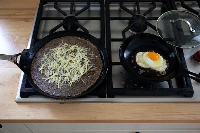 Two skillets stovetop: one holding a fried egg; one holding a crepe and some grated cheese on top.