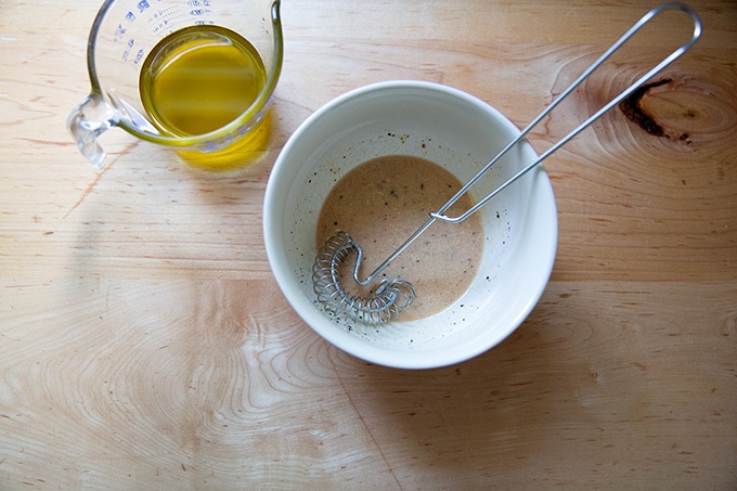 A bowl of whisked together vinegar, mustard, salt, pepper and sugar aside a liquid measure filled with olive oil.