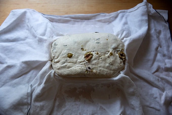 An unbaked loaf of rosemary-olive sourdough bread in a tea-towel lined banneton after 2 days in the fridge. - 45