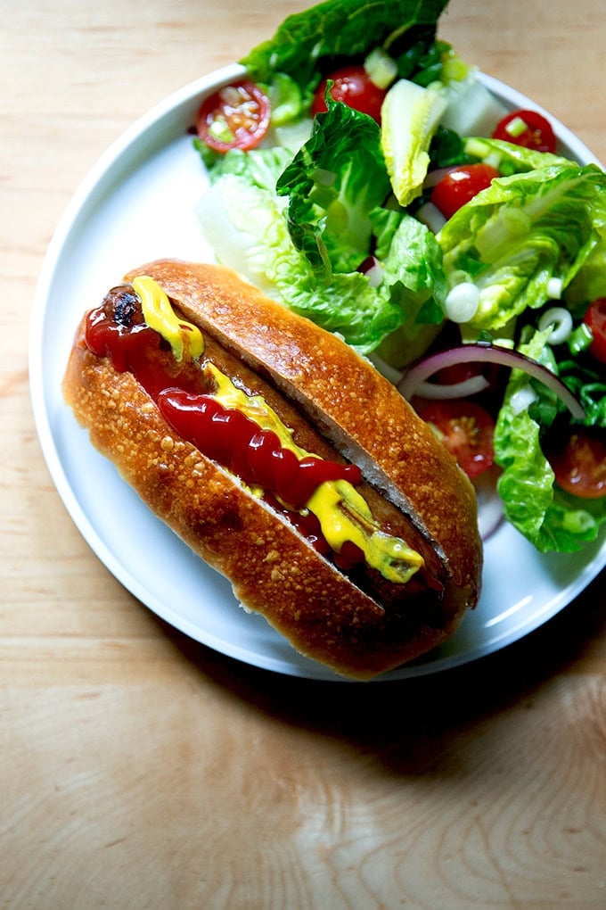 A hot dog in a brioche bun aside a Greek salad.