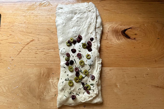 Rosemary-olive sourdough dough in the process of being folded on a countertop to incorporate the olives and rosemary. - 32