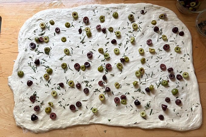 Sourdough dough stretched thinly across a work surface with olives and rosemary sprinkled across it. - 30