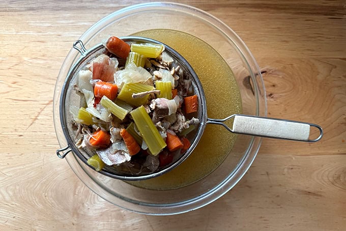 A strainer filled with boiled vegetables and chicken bones over a bowl of broth.