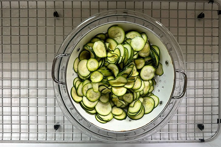 A colander filled with salted and drained zucchini set in a large glass bowl set in the sink.