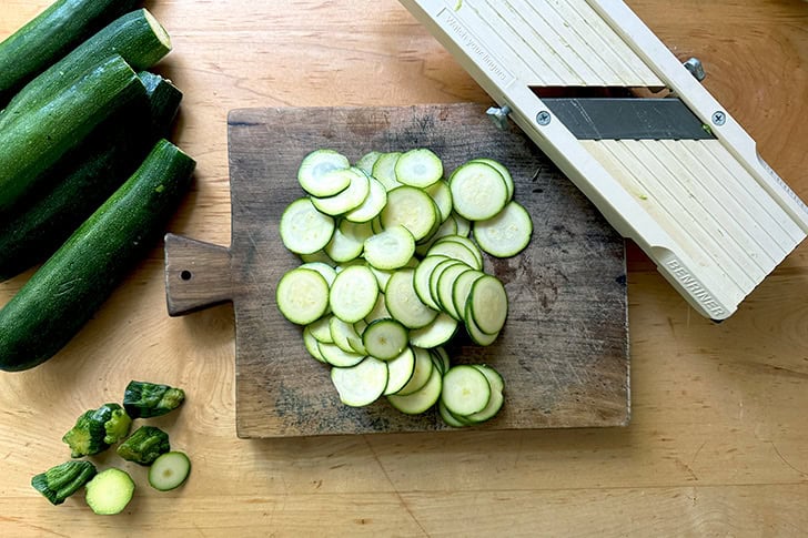 A cutting board topped with thinly sliced zucchini aside a mandoline.