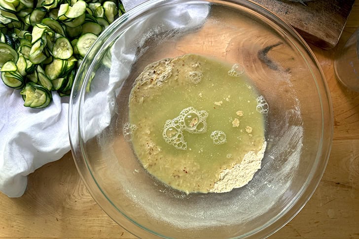 Zucchini water added to the dry ingredients to make Scarpaccia in a large glass bowl.