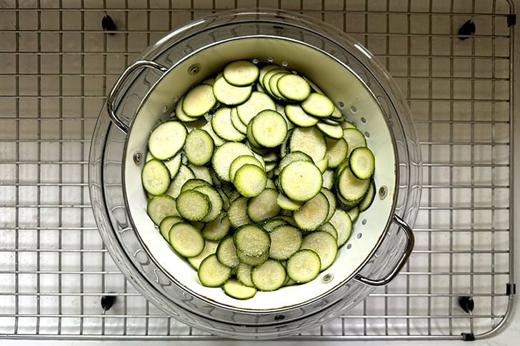 A colander filled with salted zucchini set in a large glass bowl set in the sink.